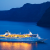 A brightly-lit cruise ship sits in a dark harbour surrounding by dark water, dark skies, and faintly visible mountains in distance