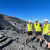 Two Leeds College of Building apprentices and their roofing lecturer stand in high-vis jackets on the edge of a quarry. The sky is amazingly blue behind them.