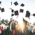 A group of professional economist apprenticeship graduates smile and laugh as they throw their board-and-mortar hats up against a blue sky