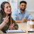 Representing Bradford 2025 creative apprentices, the image shows a young woman sat at a desk in a meeting room. She's speaking to her colleagues around the table and smiling and gesturing.