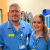 A father and daughter stand side-by-side in matching blue nurse's scrubs. The father wears glasses and a smile with his arm around his daughter. His daughter smiles and wears a lanyard too. They are both standing in the doorway to a nurses cloak room.