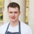Head Chef of Rudding Park’s Horto Restaurant, Callum Bowmer. Callum is a white man with short hair. He is wearing a white t-shirt and a grey apron and is smiling at the camera. We can tell he is standing against a green garden of some kind but the background is blurred.