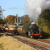A red and black steam train powers along the track through an idyllic North Yorkshire countryside scene. The train has trees either side and in the distant background there are rolling hills. The sky is a brilliant blue.