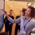 A woman in a smart shirt and lanyard points out something on a whiteboard using a whiteboard marker as her pointer. In the background and foreground a crowd of people listen to her intently as though she is presenting some information.