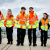 Six young engineering apprentices, male and female, walk across a walkway wearing bright yellow and orange high-vis jackets, black trousers, and heavy boots.