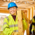 A happy young white female construction apprentice in a high-vis jacket and hard hat carries lumber on a building site. She smiles into the camera.