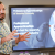 A professional man stands at the front of a classroom. He is gesticulating and wearing a jazzy shirt. Behind him a TV screen shows he is presenting on the topic of professional apprenticeships at Teeside University
