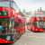 Two red London buses loom over the camera as they zip around London streets. A grand building stands in the background.