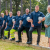 A group of men in navy polo shirts stand in a wooded area. All of them have one from on a raised bed and one foot towards the back. They have all placed their left hand on the raised leg. They are all smiling. At the end closest to the camera a women is smiling and wearing a blue dress.