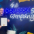 A man stands holding a certificate and smiling in front of a large neon sign that says "The Sussex Sign Company"